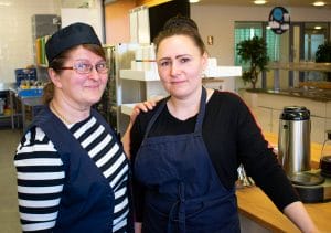 Kaksi iloista naista hymyilee kameralle kahvilinjaston edessä