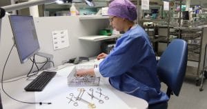 Sairaalan suojavaatteisiin pukeutunut nainen tarkastaa toimenpiteissä ja leikkauksissa käytettäviä välineitä työpöydän ääressä.