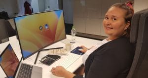 Nainen istuu työpöydän ääressä, jossa on tietokone ja hymyilee.