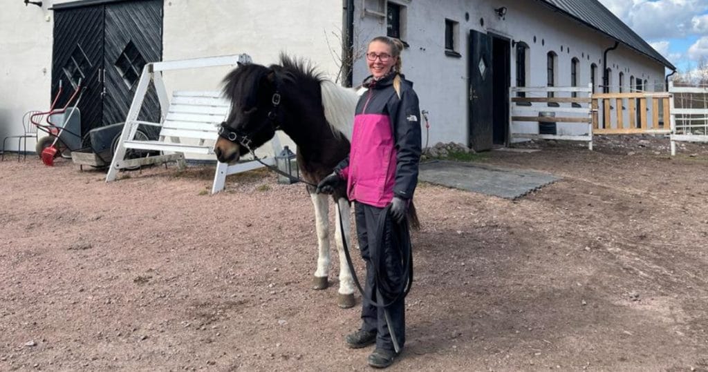 Nainen seisoo islanninhevosen vieressä hevostallin ulkopuolella.
